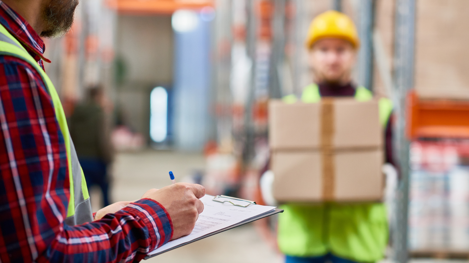 Worker holding cardboard boxes while another checks a clipboard in a warehouse, wearing safety vests and helmet.