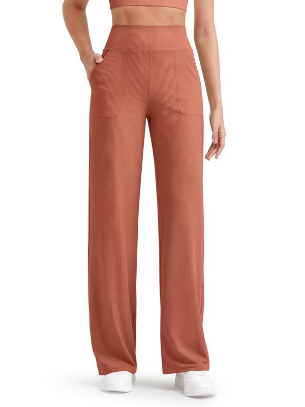 A pair of high-waisted ribbed yoga pants in rust with side pockets, worn by a model with white sneakers, highlighting the relaxed fit and textured fabric