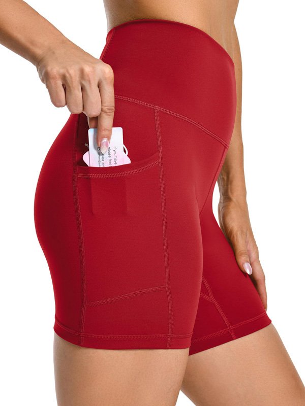 Woman wearing high-waisted red athletic shorts, placing a card into the side pocket, showcasing a smooth, stretchy fit and practical design