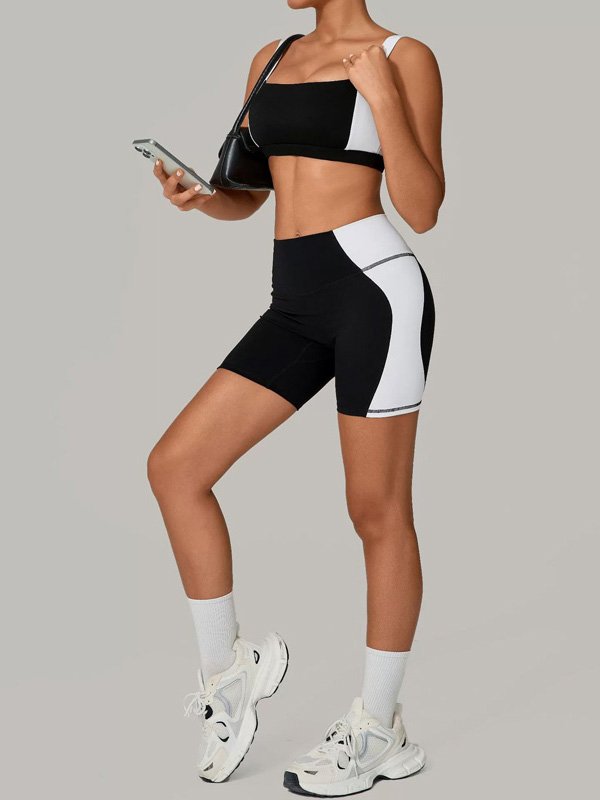 Woman wearing a black and white color-block athletic set with matching bike shorts and crop top, holding a phone, paired with white sneakers and socks, in a relaxed, sporty pose.