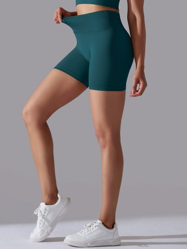 Woman wearing high-waisted teal athletic shorts, adjusting the waistband, paired with white sneakers, highlighting the smooth, stretchy fit.