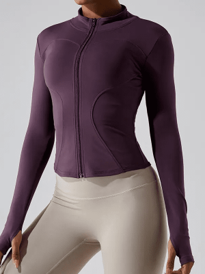 Woman wearing a fitted purple zip-up activewear jacket with thumbholes, paired with beige high-waisted leggings.